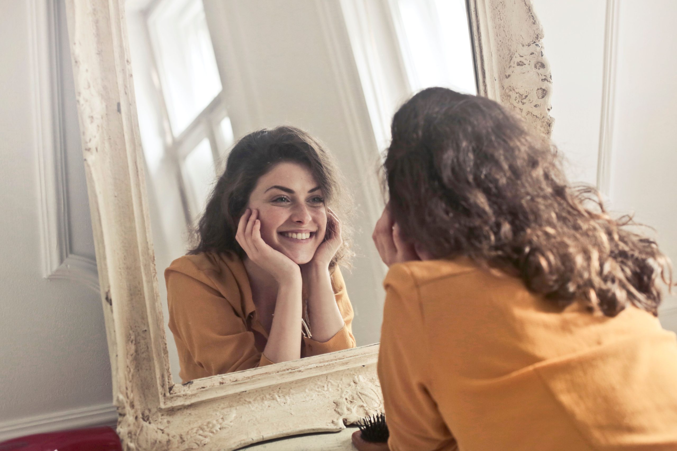 A cheerful woman smiling warmly at her reflection in a vintage mirror, embracing her genuine self-worth